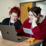 Two people sat together at a table both looking at the same laptop
