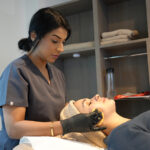 A lady with black hair, grey uniform and black gloves with her hands on a clients face who's laying on a treatment bed