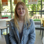 A female with long blonde hair and a light blue denim jacket sat at a table, a wall of windows in the background