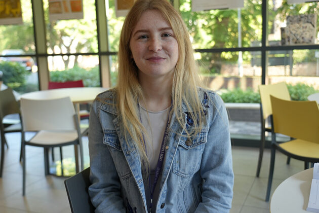A female with long blonde hair and a light blue denim jacket sat at a table, a wall of windows in the background