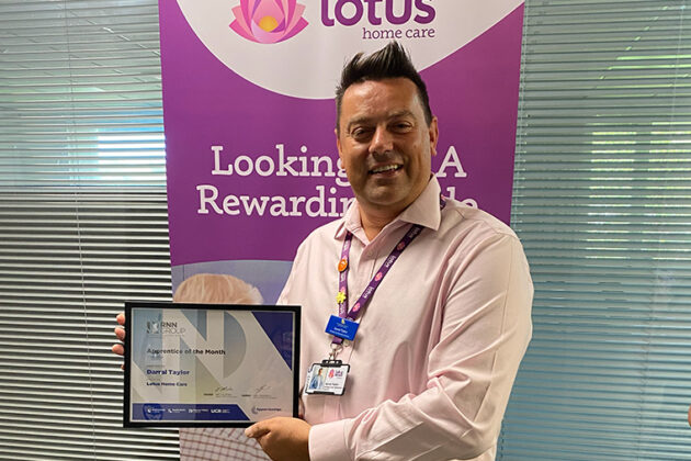 A smiling male holding a framed certificate, a pull up banner behind him that says Lotus