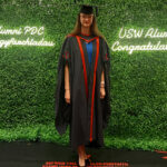 Hannah stood in her graduation attire in front of a green hedge with lit up words 'USW Alumni Congratulations'