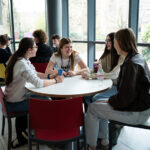 Two tables each full of smiling students socialising, large windows behind the tables